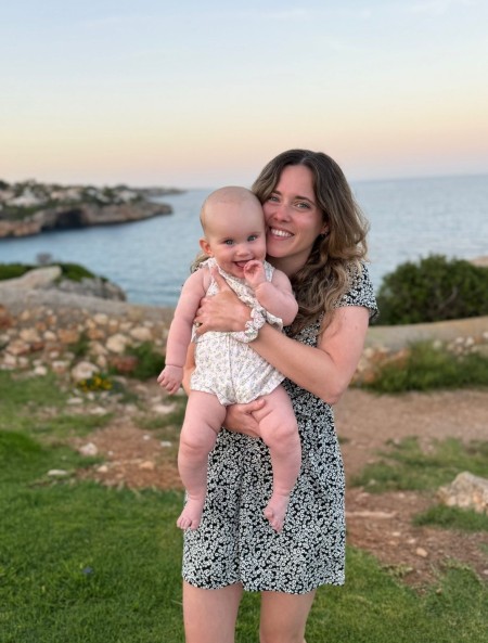  A person standing outdoors near a rocky coastline at sunset, holding a small child. The person is wearing a short patterned dress, and the child is dressed in a light-colored outfit. The sea and distant shoreline are visible in the background, with soft pastel sky colors above.
