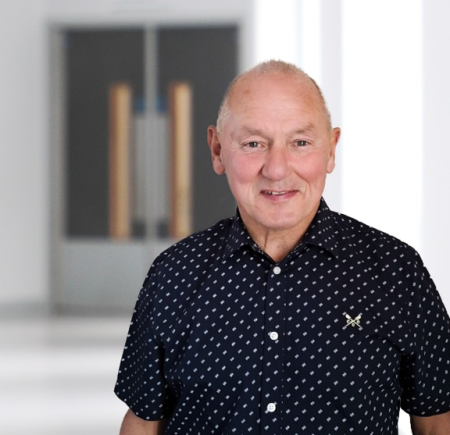  Smiling man standing indoors in front of double doors. He has short gray hair and is wearing a navy blue short-sleeved shirt with a small white geometric pattern.