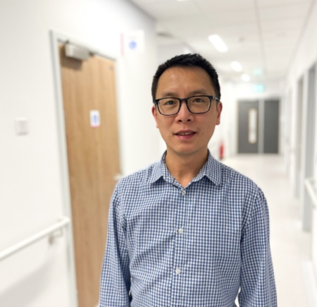 Man wearing glasses and a blue checkered shirt standing in a bright, modern hallway with white walls and doors.