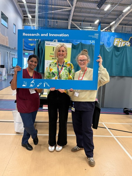Three people stand inside a blue University Hospitals of Liverpool Group Research and Innovation photo frame at a community event venue. Research and information stands are visible in the background, including an NIHR BioResource display.