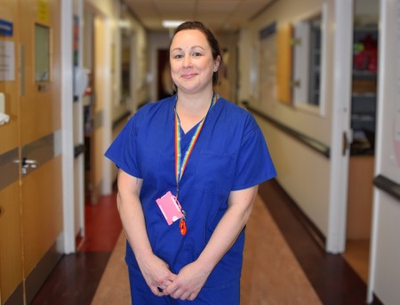 Photo of Kat Davies, Consultant Head and Neck Surgeon, at Aintree University Hospital, in a hospital corridor in blue scrubs.