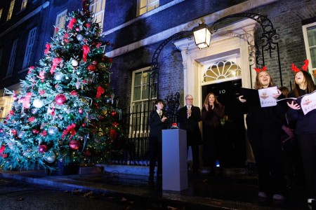 Prime Minister Keir Stramer, standing in front of a podium with a red button on top. To his right is a large Christmas tree decorated with red ribbons and baubles and to his left a choir wearing red reindeer antlers. 