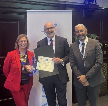 Three individuals standing and smiling at an award ceremony. Professor Terry Jones holds a certificate and trophy after receiving the ENT UK Lifetime Trainer Award 2025. A woman in a red suit and a man in a dark suit stand beside him.